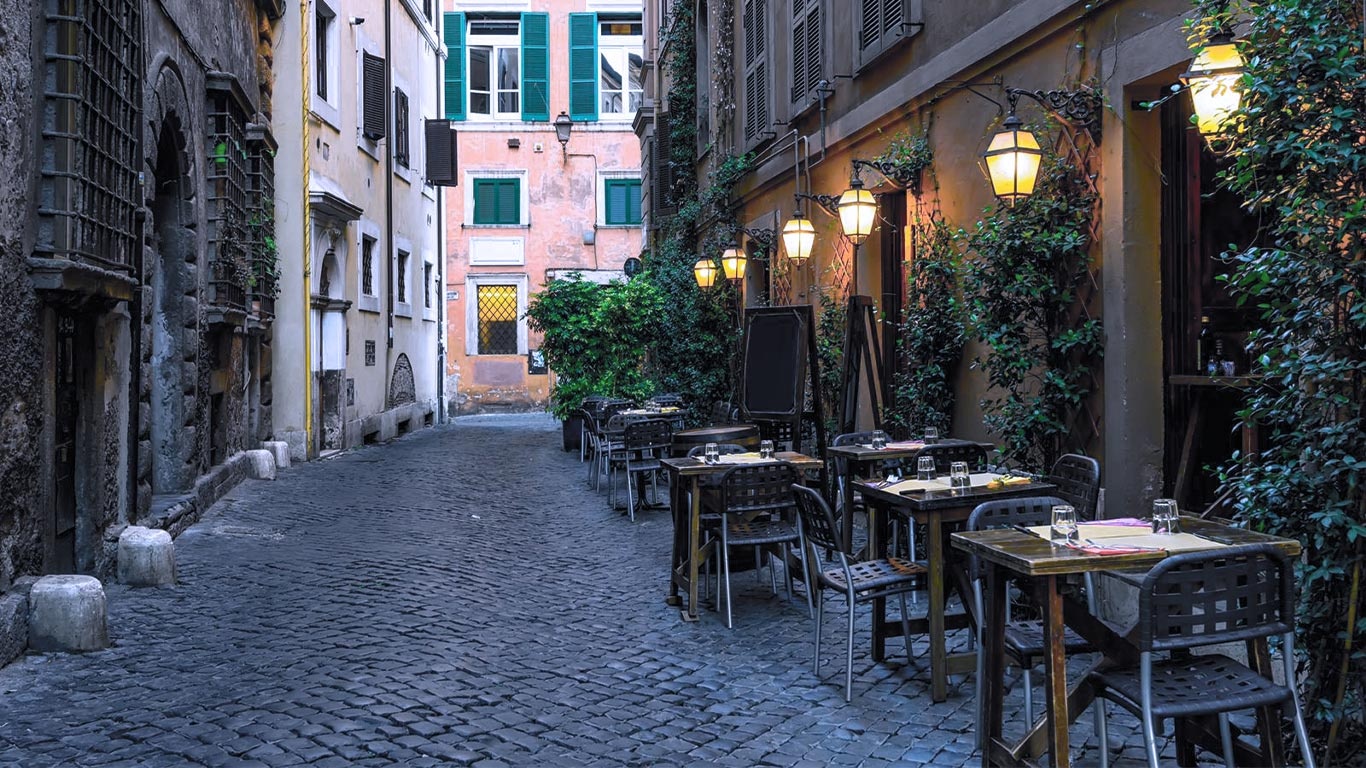 A narrow cobbled street in Trastevere lined with outdoor tables and lanterns